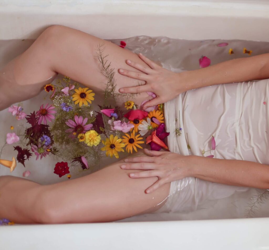 pernas de mulher aberta deitada em uma banheira cheia de água e pétalas de diversas flores