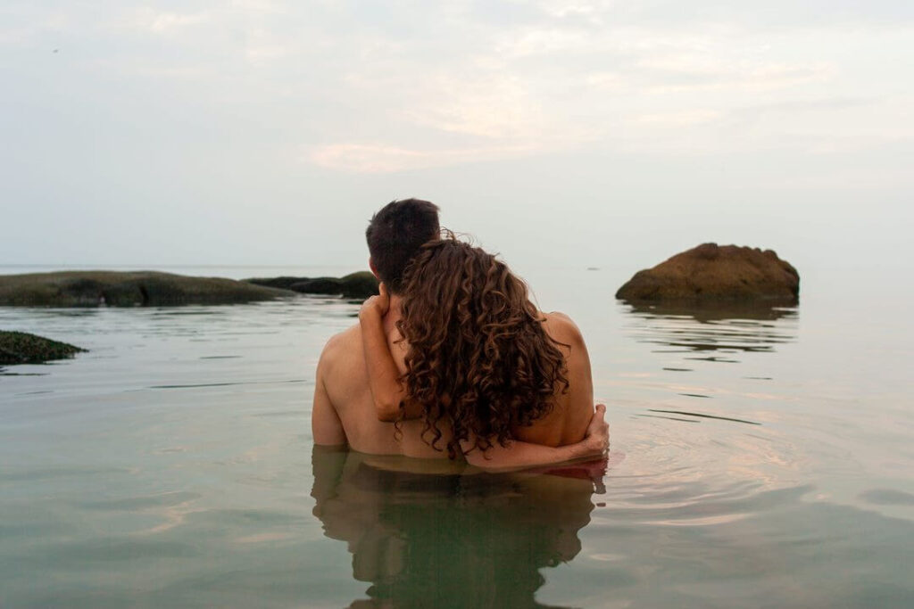 um homem e uma mulher estão abraço dentro do mar. Os dois estão de costas e aparentar estarem pelados