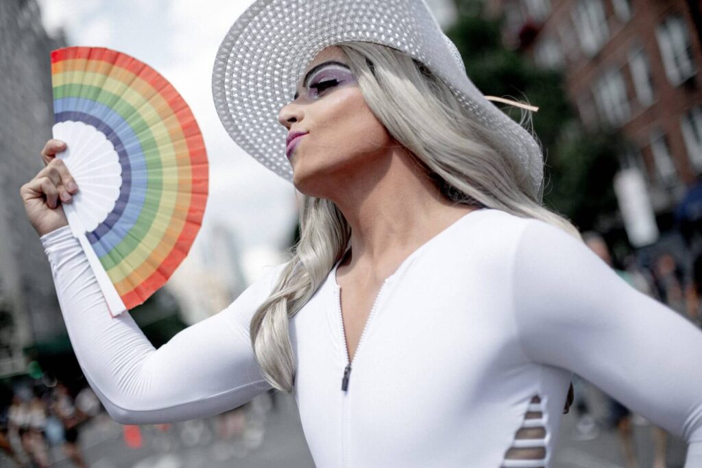 uma drag queen está de perfil abanando um leque com as cores da bandeira LGBTQIA+
