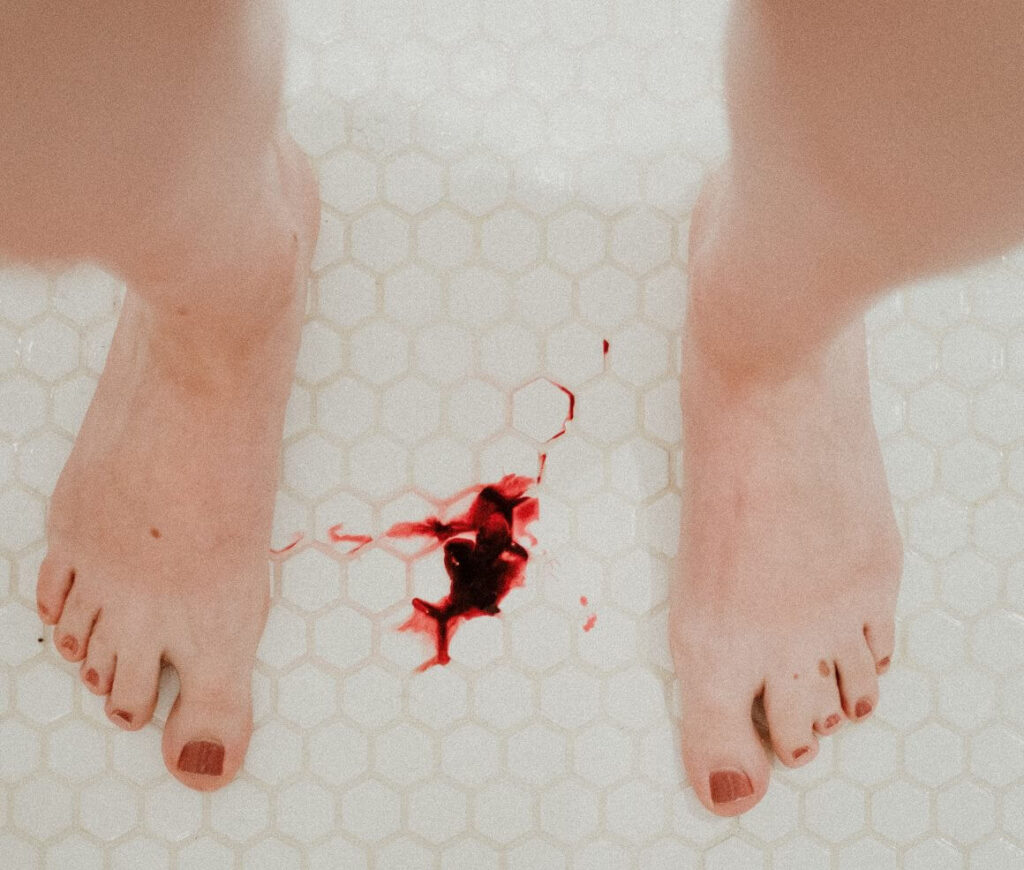 foto dos pés de uma mulher no banheiro. Entre seus pés, no chão, tem uma mancha de sangue
