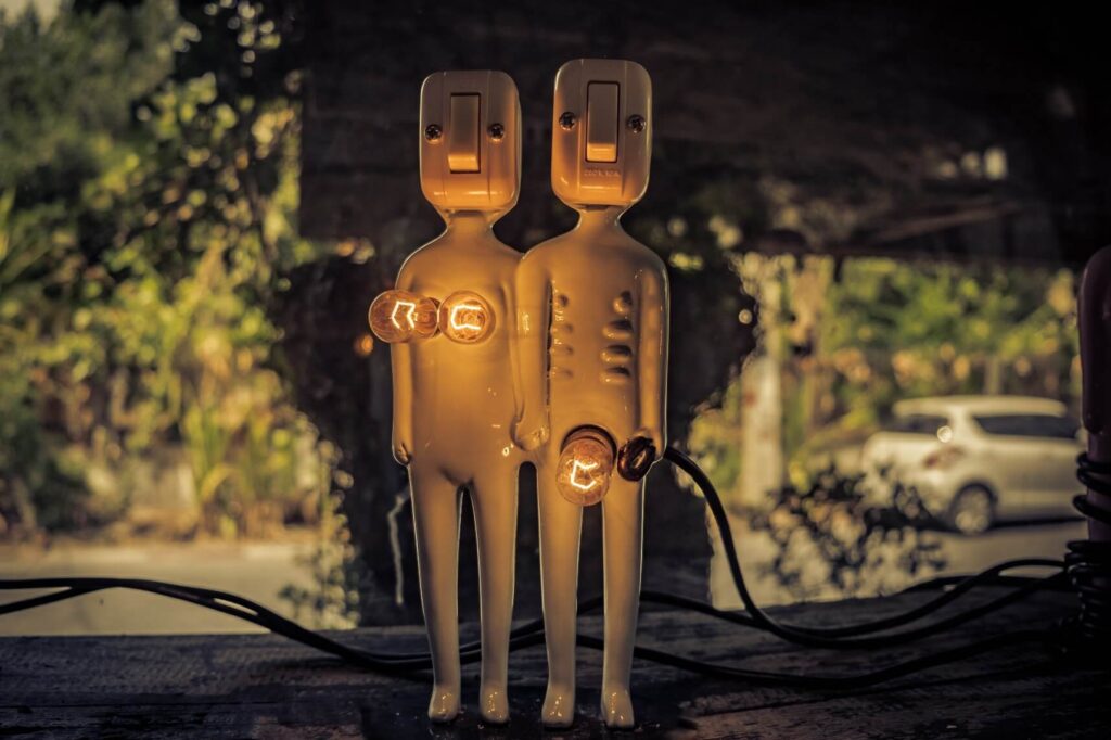 foto de dois bonecos que representam um homem e uma mulher. Os rostos do bonecos são feitos com interruptor de luz e o boneco que representa a mulher possui duas lâmpadas acessas onde seriam os seios e o boneco masculino uma luz acesa onde seria o pênis