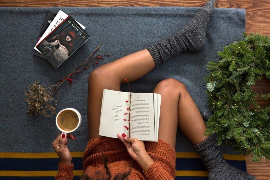 mulher sentada no chão lendo um livro e segurando uma xícara.Ao seu redor, há alguns livros e plantas ao seu lado.