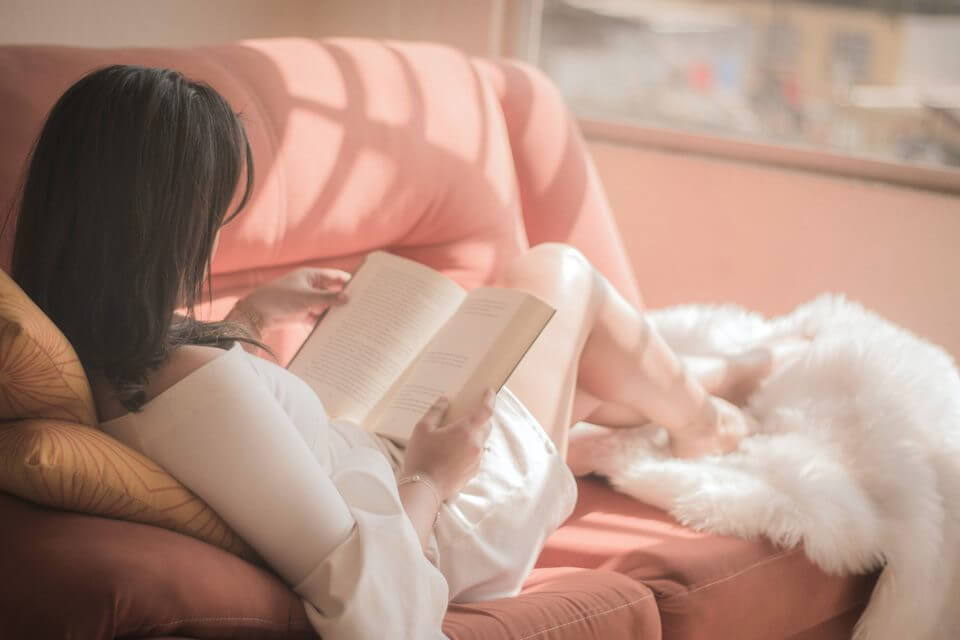 mulher sentada em um sofá lendo um livro. O local onde ela está lendo tem muita luz natural do sol. 