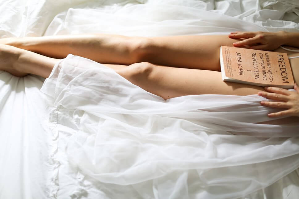 pernas nuas de mulher aparentemente sentada em uma cama. Há um livro em cima das pernas da mulher.