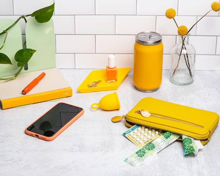 mesa com um coletor menstrual em meio a vários objetos (celular, maquiagem, carteira, esmalte, caderno e chave).