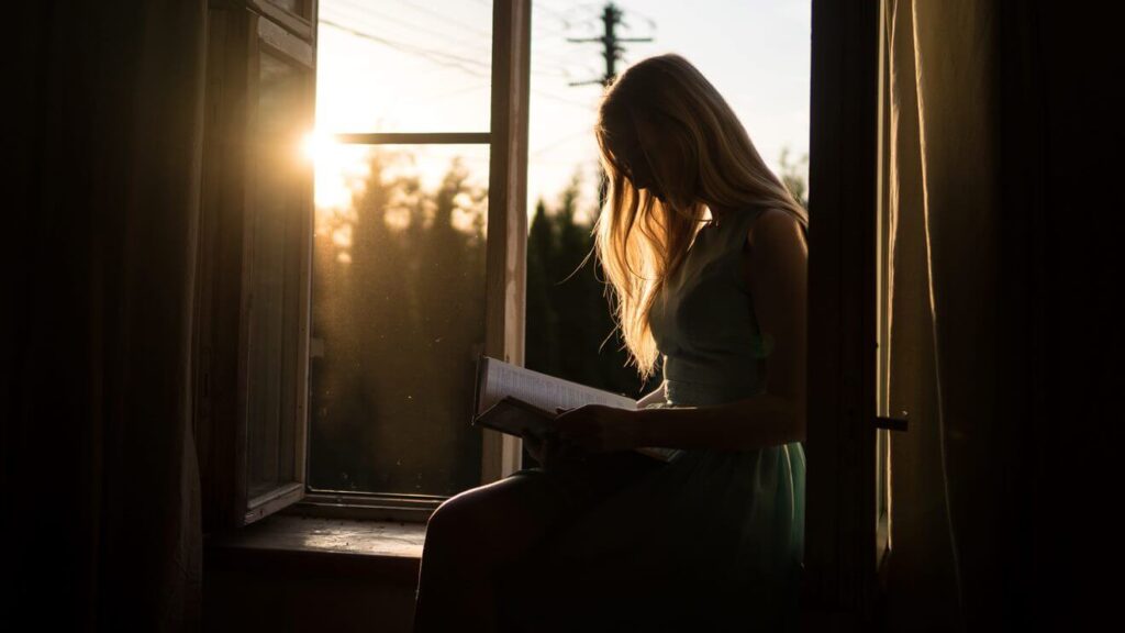 Mulher sentada em uma janela, lendo um livro ao pôr do sol.