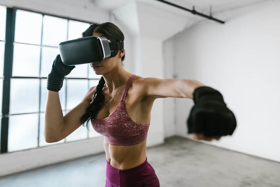 mulher está com roupa de ginástica e com óculos de realidade virtual. A mulher está em posição de luta como se estivesse fazendo exercício com o auxílio da realidade virtual 