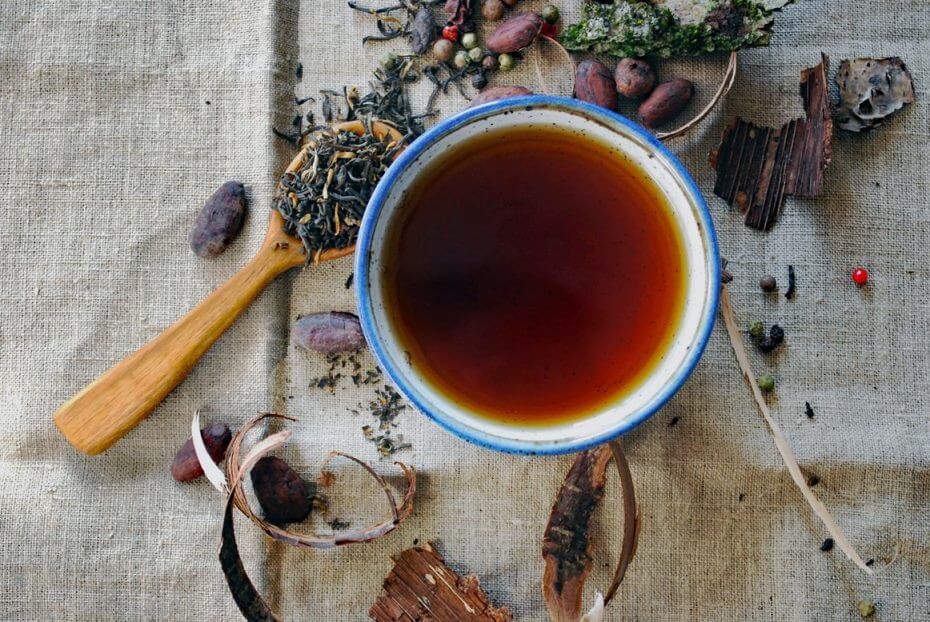 xícara cheia de chá está em cima de uma mesa com diversas ervas ao redor 