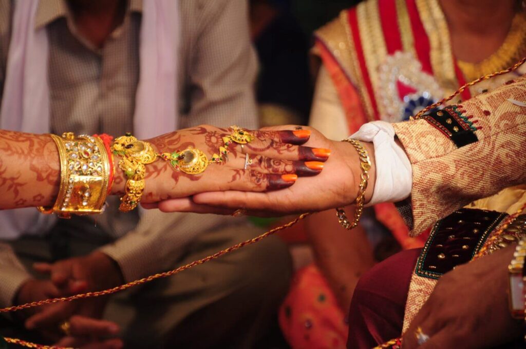 No primeiro plano da foto, há o antebraço de uma mulher e de um homem no que aparenta ser um casamento indiano. A mão da mulher está com tatuagem de henna indiana. No fundo da foto, há um homem e uma mulher sentados com roupa típica da Índia. 