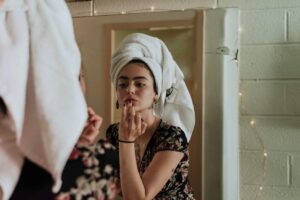 mulher com uma toalha enrolada no cabelo em frente a um espelho. A mulher está passando batom com o dedo anelar no lábio inferior.