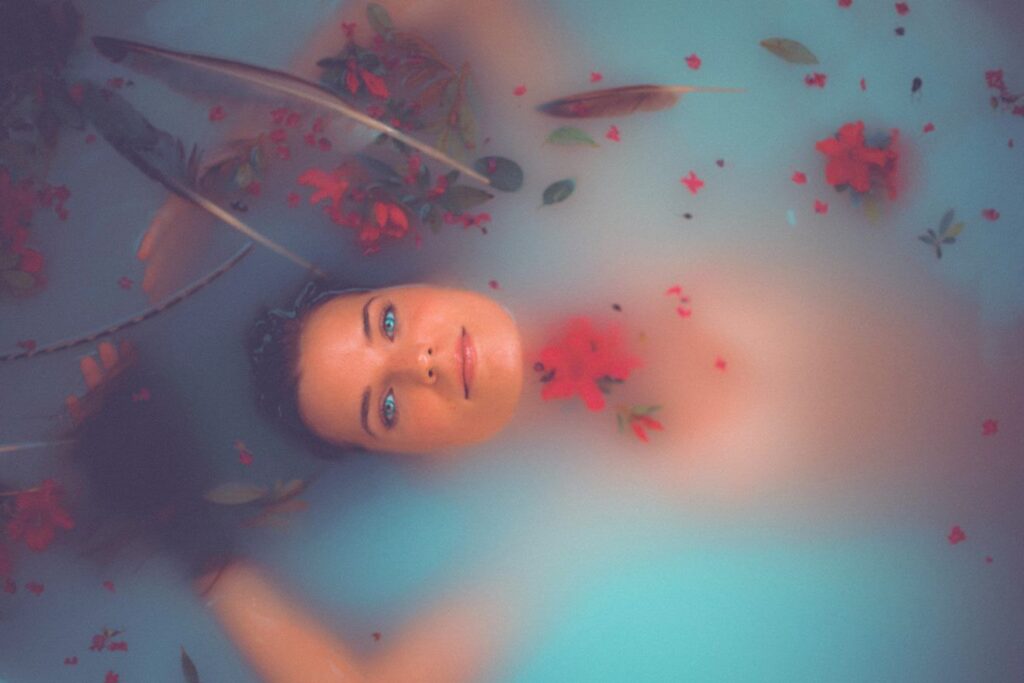 Mulher está submersa em água com aspecto turvo. A mulher está só com a cabeça na superfície e a água contém algumas flores, folhas e penas.