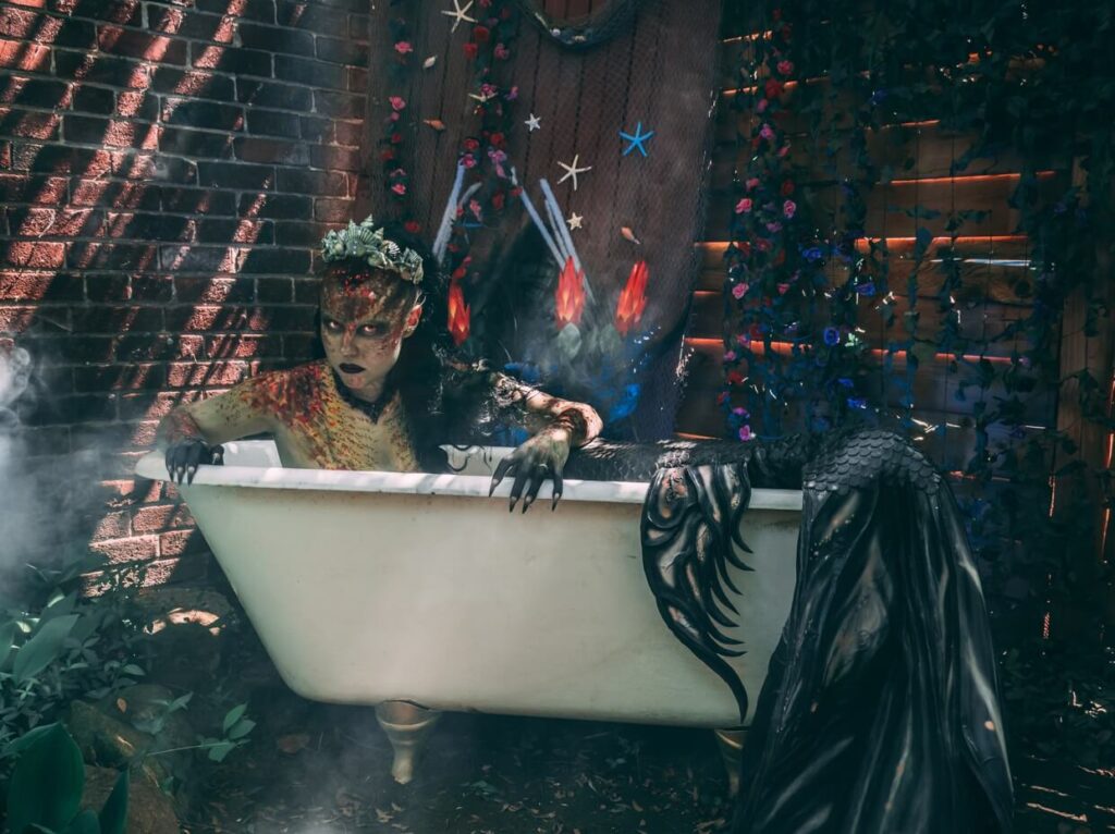 uma mulher vestida de sereia está dentro de uma banheira. A mulher olha fixamente para a câmera com um semblante sombrio