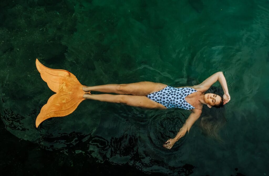 mulher de maiô está boiando em água esverdeada. A mulher está com um semblante sereno e nos seus pés há uma nadadeira em formato de calda de sereia