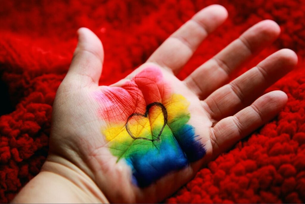 foto da palma da mão de uma pessoa. As cores da bandeira do orgulho gay estão pintadas na palma da mão e também há um coração pintado no meio da mesma palma.