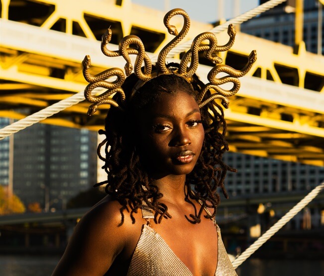 mulher muito bonita olhando diretamente para a câmera. Ela está com um vestido de alça e um arquinho na cabeça que imita as cobras que são os "cabelos" da Medusa.