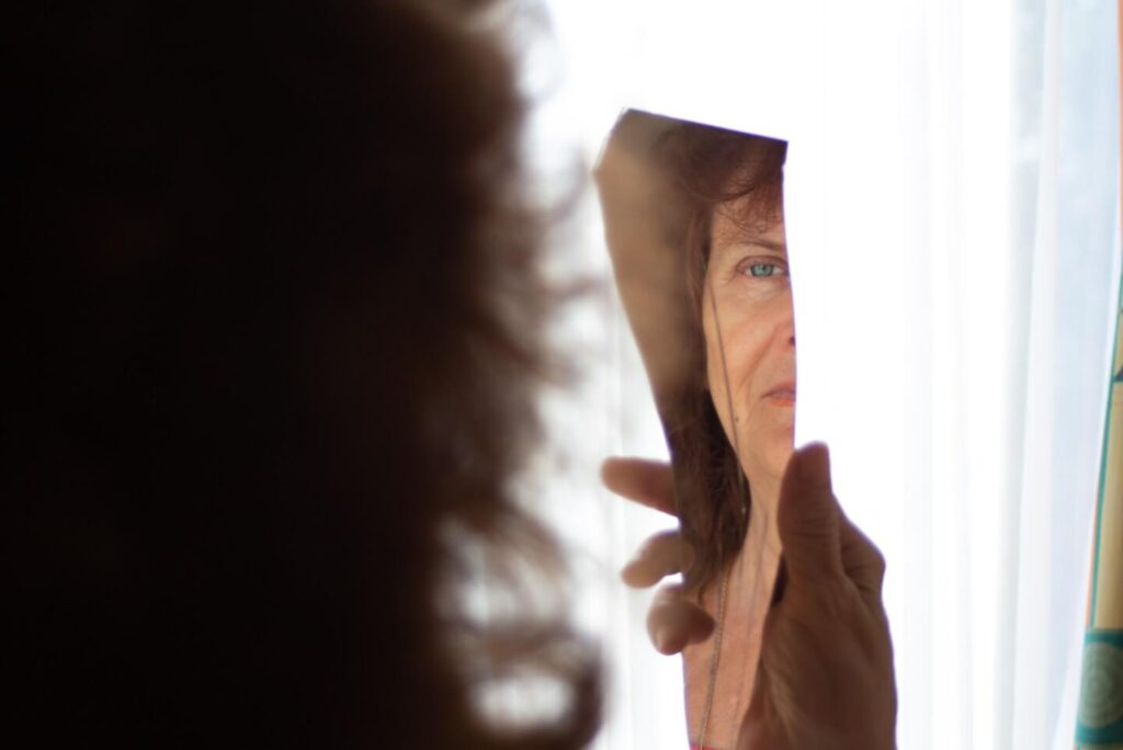 mulher está de costas segurando um espelho quebrado na altura do seu rosto. O pedaço do espelho reflete o rosto de uma mulher idosa.